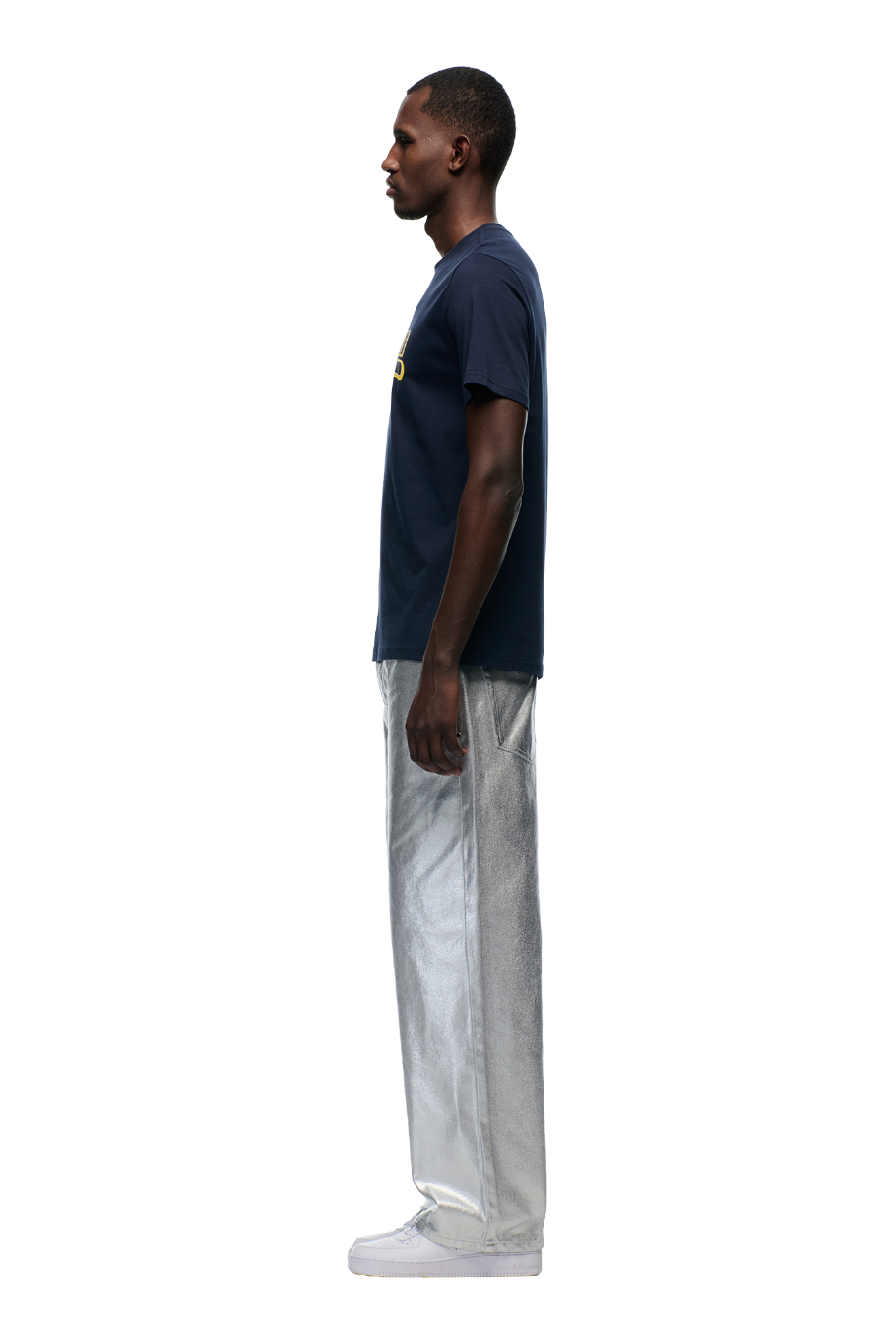 A man stands in profile against a plain background, wearing the 6PM CLEAN EDGE WAVE T-SHIRT MIDNIGHT BLUE, paired with shiny metallic silver pants and white shoes.