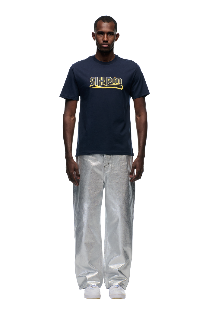 A man with short hair stands facing forward in a 6PM CLEAN EDGE WAVE T-SHIRT MIDNIGHT BLUE, featuring yellow text, paired with wide metallic silver pants and white sneakers, set against a plain white background.