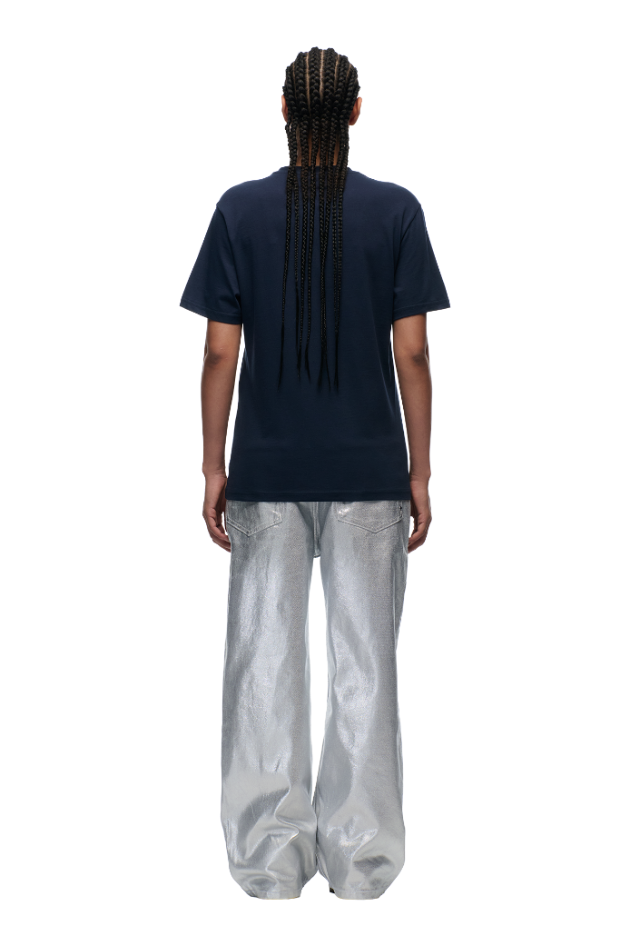 A person with long braids faces away, wearing the 6PM CLEAN EDGE WAVE T-SHIRT in Midnight Blue paired with shiny, loose-fitting silver pants against a plain white background.