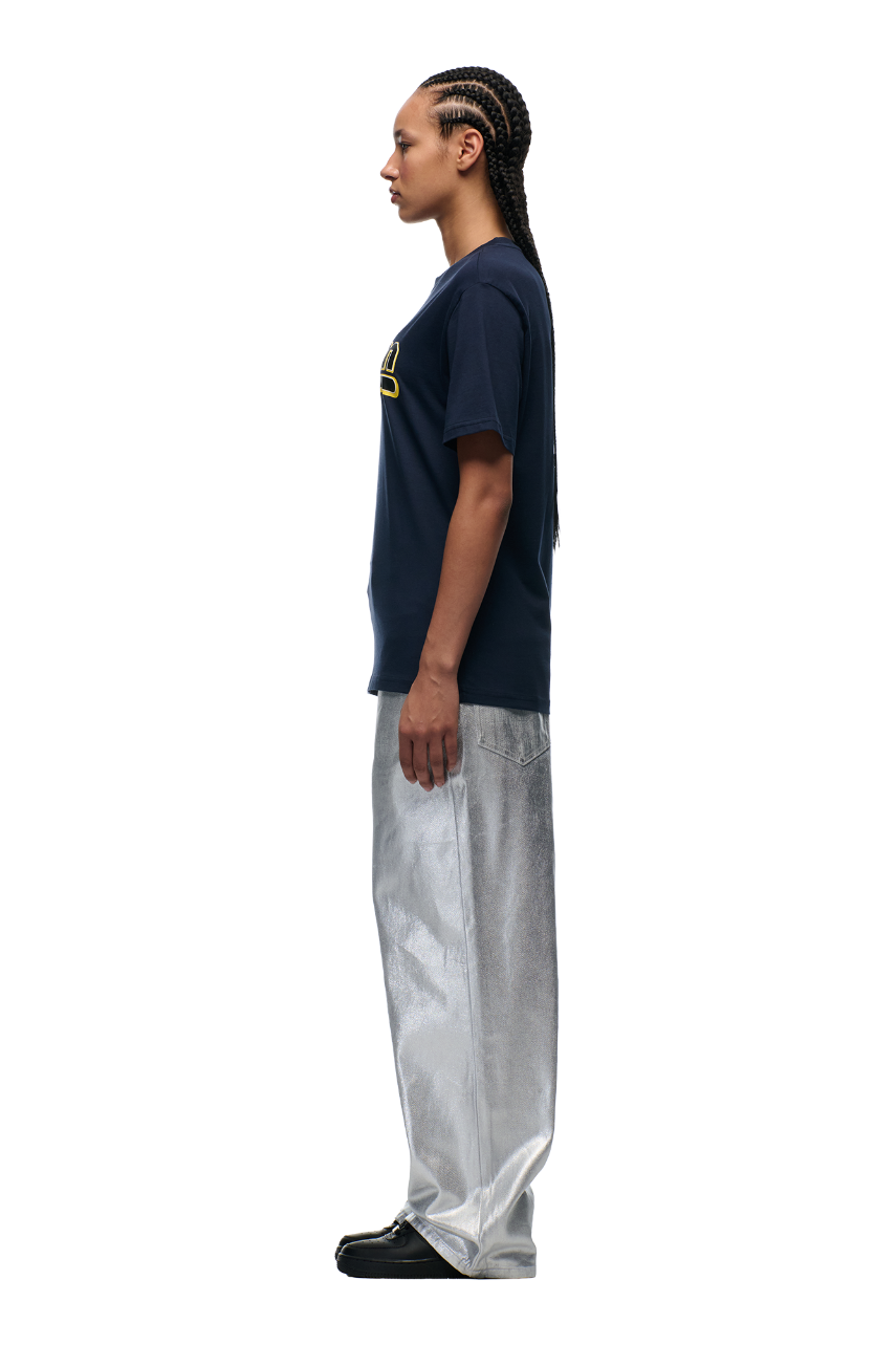 A person with long braided hair stands in profile against a plain background, wearing the 6PM CLEAN EDGE WAVE T-SHIRT MIDNIGHT BLUE, loose silver metallic pants, and black shoes.