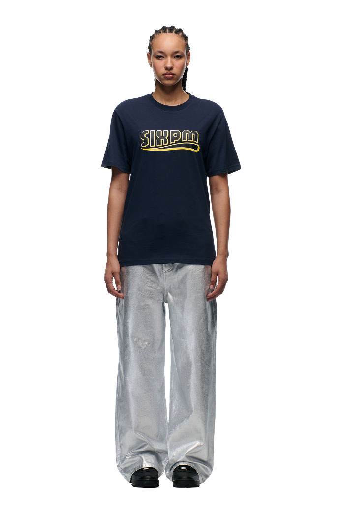 A person with braided hair wears a midnight blue CLEAN EDGE WAVE T-SHIRT by 6PM, loose silver metallic pants, and black shoes, standing facing forward against a plain white background.
