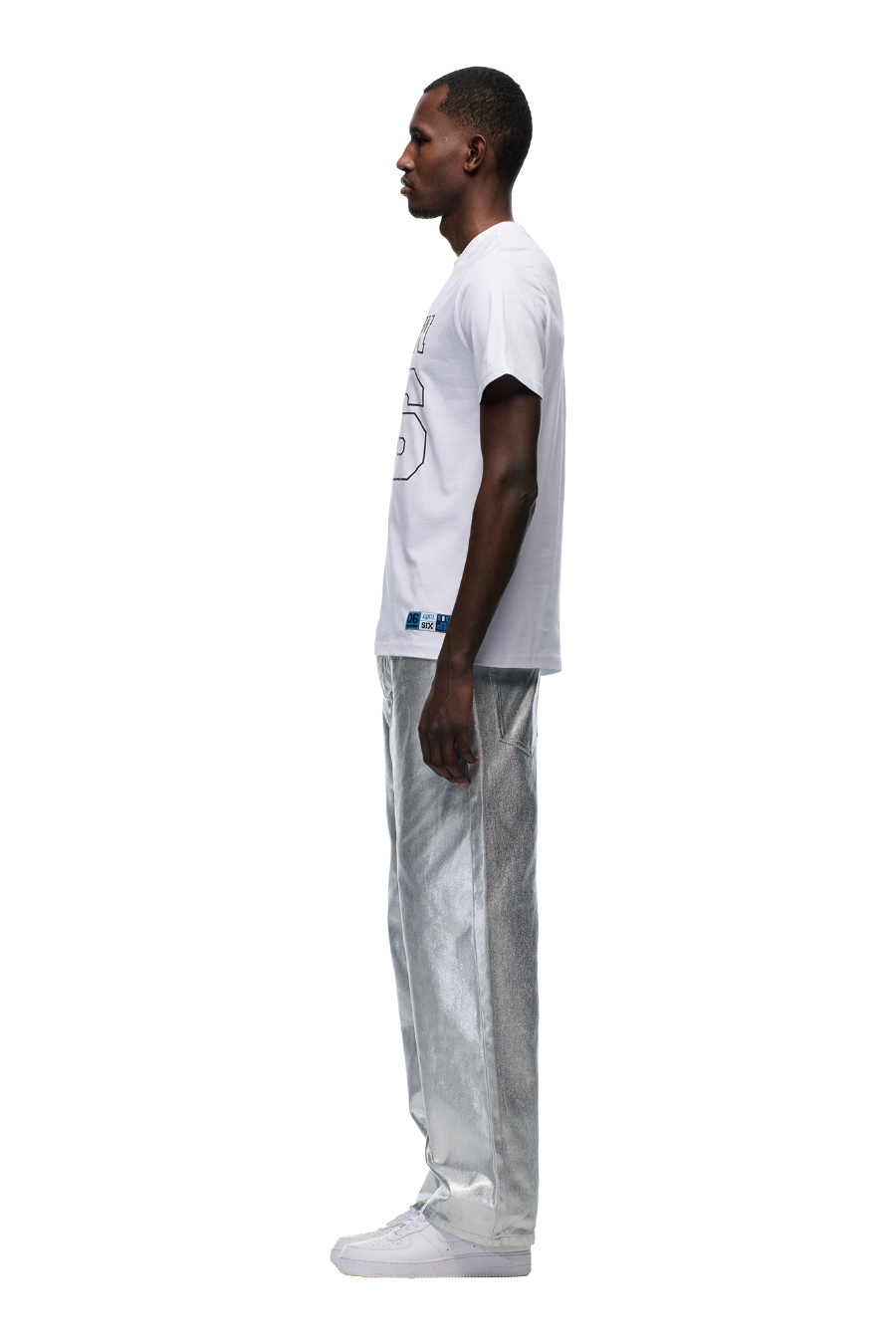 A man stands in profile against a white background, wearing the 6PM DOWN UNDER COLLEGE T-SHIRT WHITE, paired with shiny metallic silver pants and white sneakers.