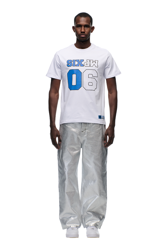 A man stands facing forward wearing the 6PM DOWN UNDER COLLEGE T-SHIRT in white, paired with shiny silver wide-leg pants and white sneakers, set against a plain white background.