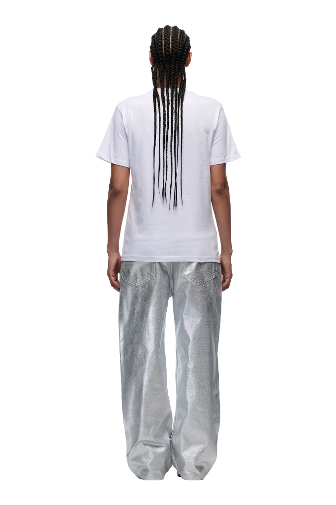 A person with long braids stands facing away in a 6PM DOWN UNDER COLLEGE T-SHIRT WHITE, featuring a woven side label, paired with loose shiny silver pants. The background is light and plain.