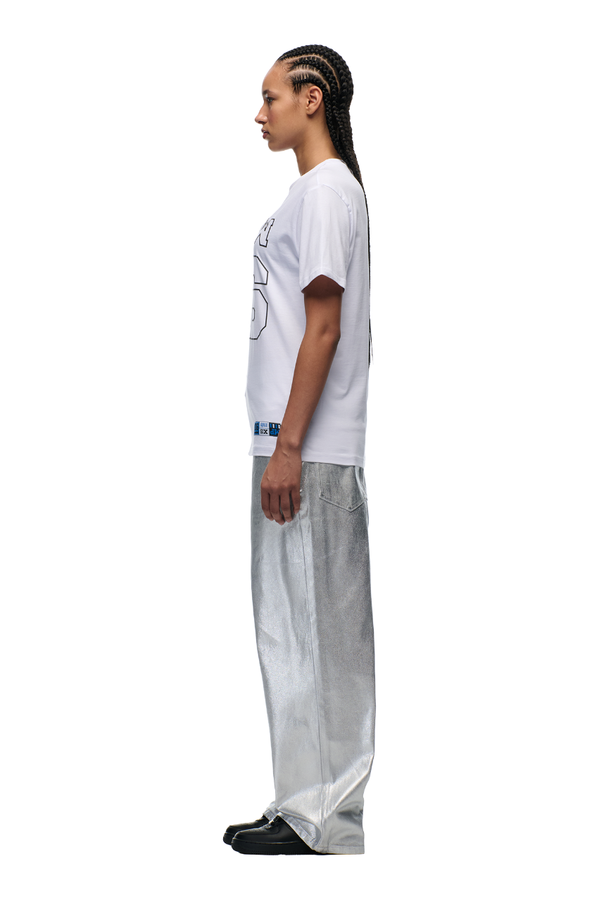 A person with long braids stands in profile against a plain background, wearing the 6PM DOWN UNDER COLLEGE T-SHIRT WHITE with a high-density print, paired with loose silver metallic pants and black shoes.