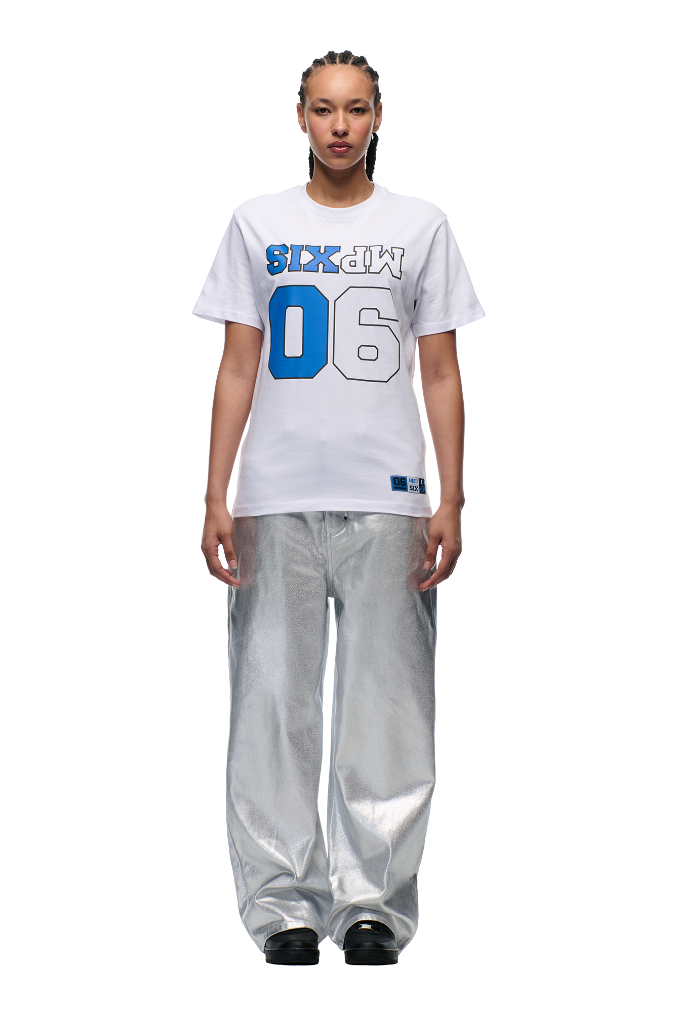 A person faces forward wearing the 6PM DOWN UNDER COLLEGE T-SHIRT WHITE with blue "06" and mirrored "M PXIS" print, paired with shiny silver wide-leg pants and black shoes, set against a plain background.