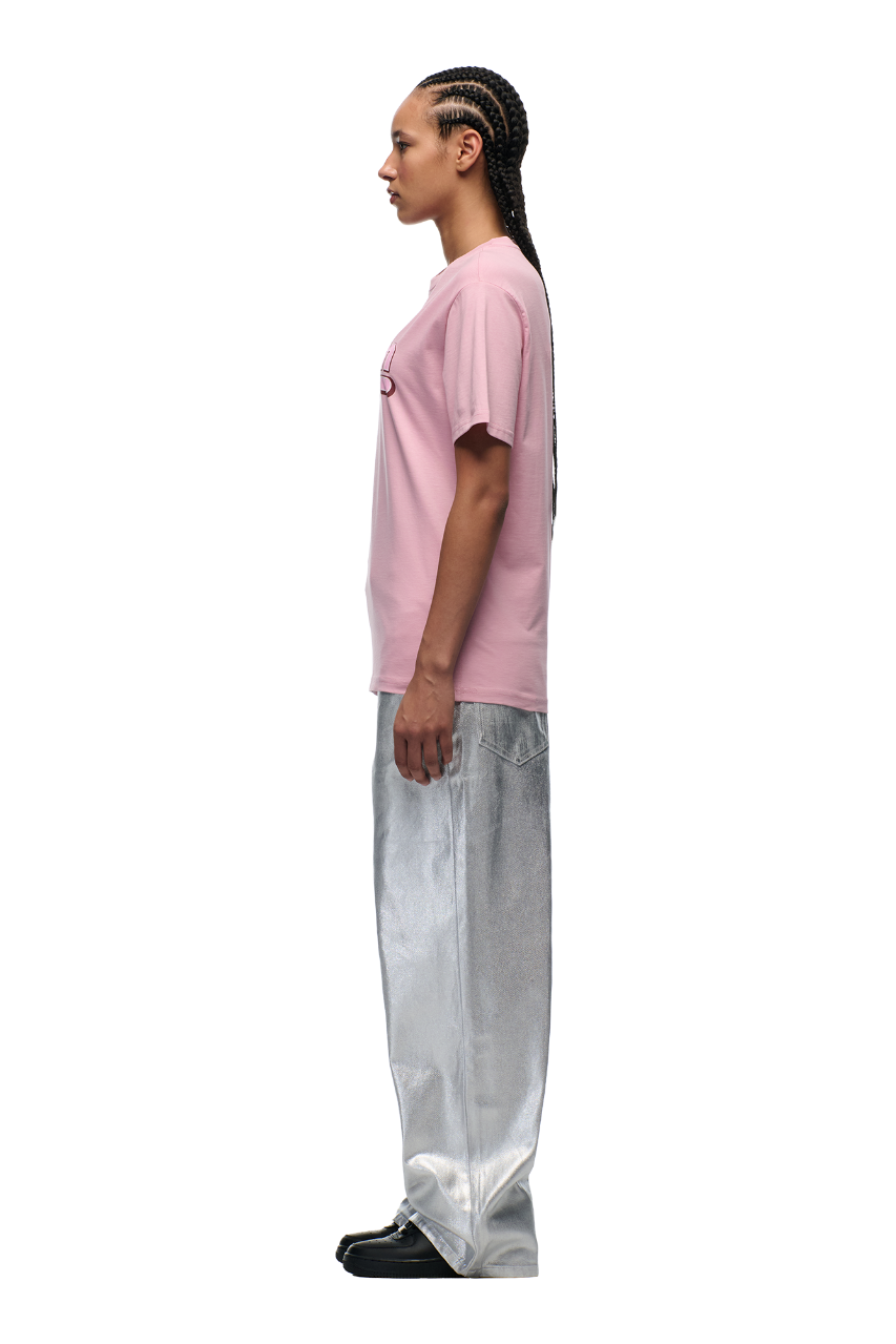 A person with long braided hair stands in profile, wearing the 6PM CLEAN EDGE WAVE T-SHIRT PINK, shiny silver wide-leg pants, and black shoes against a plain white background.