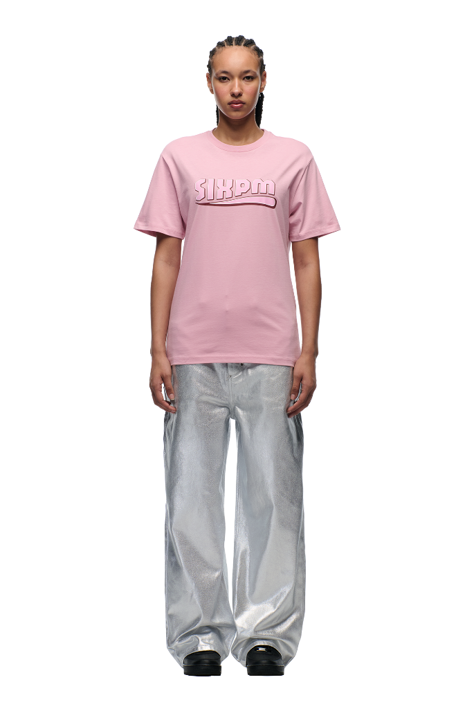A person with braided hair wears the 6PM CLEAN EDGE WAVE T-SHIRT PINK and loose, shiny silver pants, standing against a plain white background.