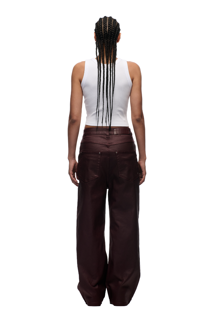 A person with long braids, in a white tank top and 6PM WAX COATED DENIM DARK BURGUNDY pants with matte silver details, stands facing away against a plain white background.