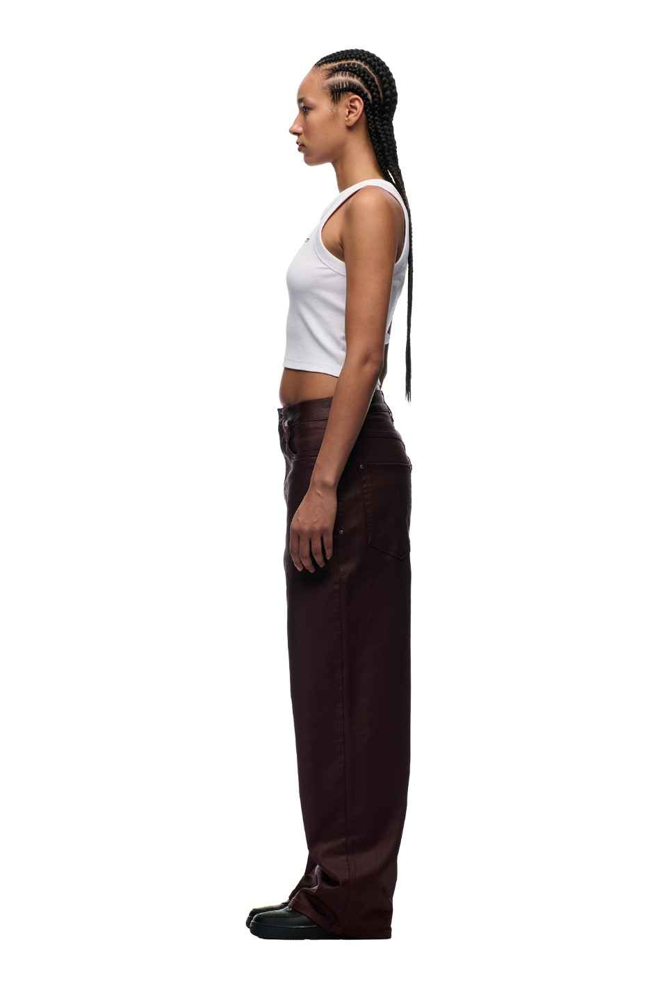 Woman with long cornrow braids stands in profile, wearing a white sleeveless crop top, 6PM WAX COATED DENIM DARK BURGUNDY pants with matte silver details, and black shoes, against a plain white background.