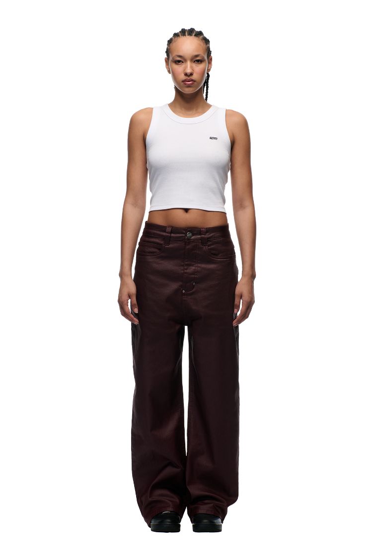 A person faces forward against a plain white background, wearing a white cropped tank and 6PM's WAX COATED DENIM DARK BURGUNDY pants paired with black shoes.