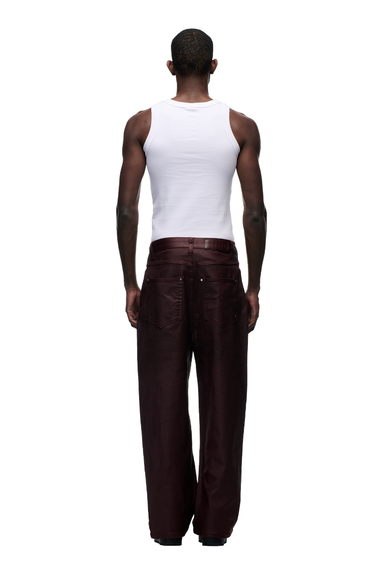 A person stands with their back to the camera in a white sleeveless shirt and 6PM’s WAX COATED DENIM DARK BURGUNDY pants featuring matte silver details, set against a plain light gray background.