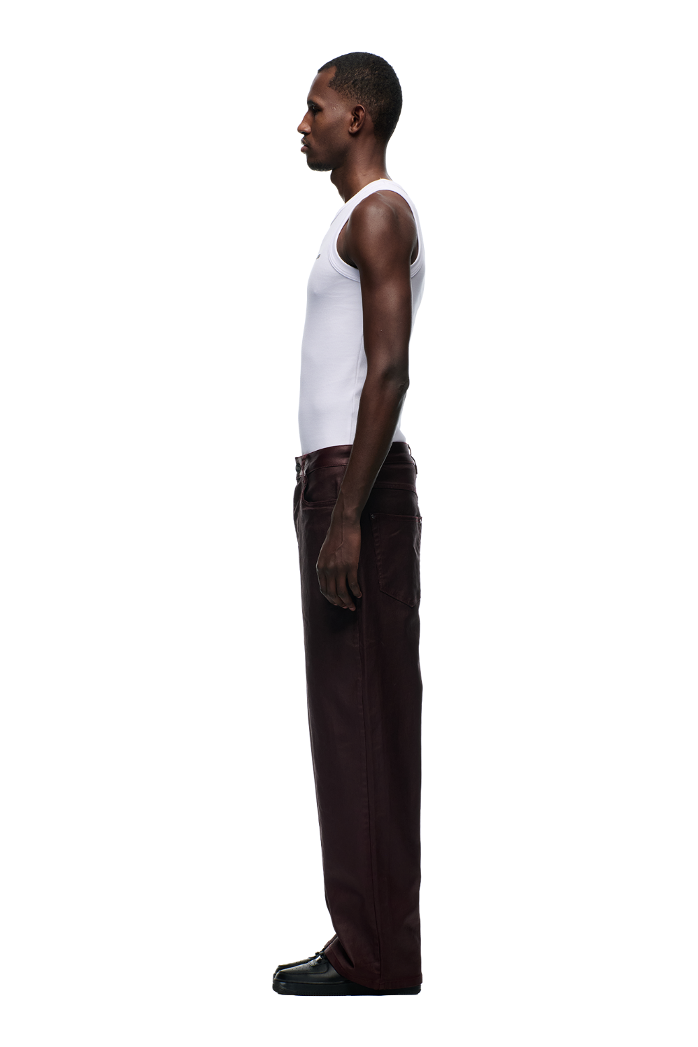 A man stands in profile against a plain background, wearing a white sleeveless shirt, 6PM WAX COATED DENIM DARK BURGUNDY pants with matte silver details, and black shoes.