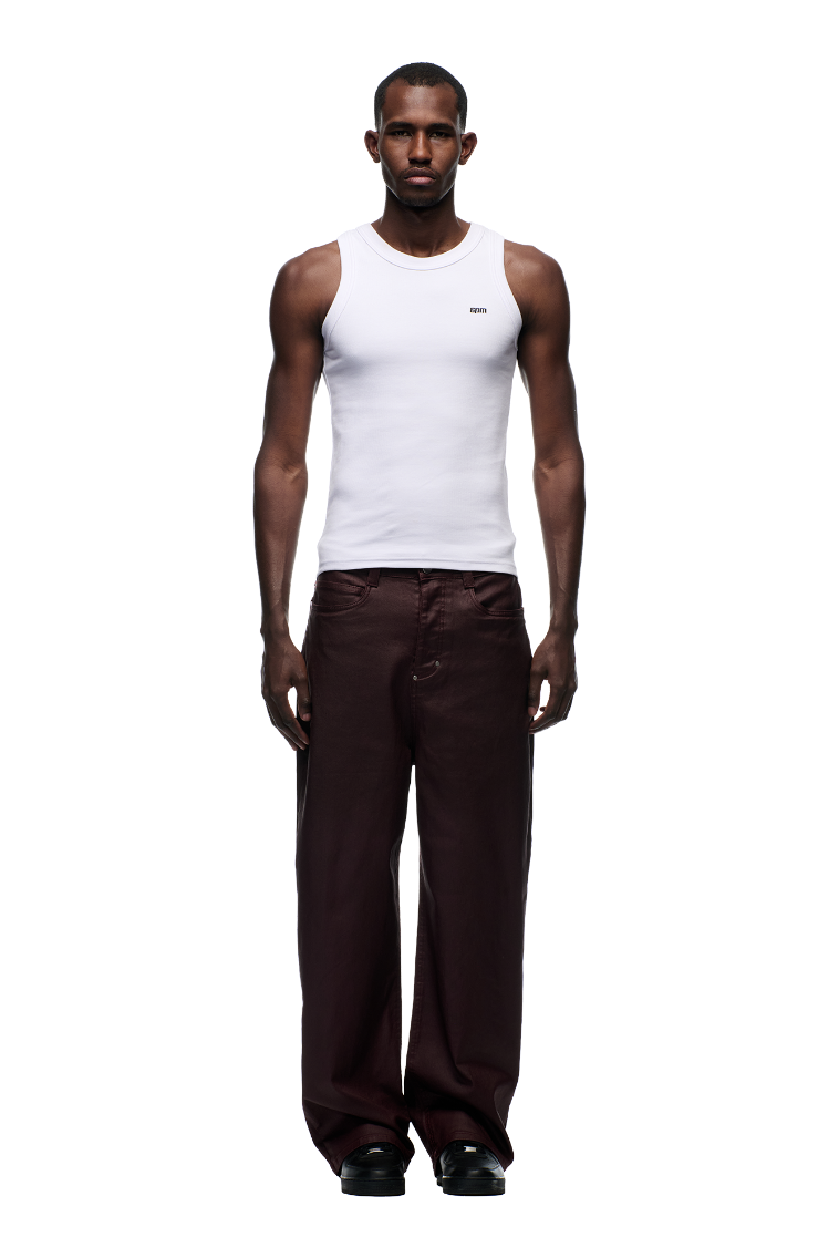 A man faces forward against a white background, wearing a white sleeveless tank top, black shoes, and 6PM WAX COATED DENIM DARK BURGUNDY pants with matte silver details.