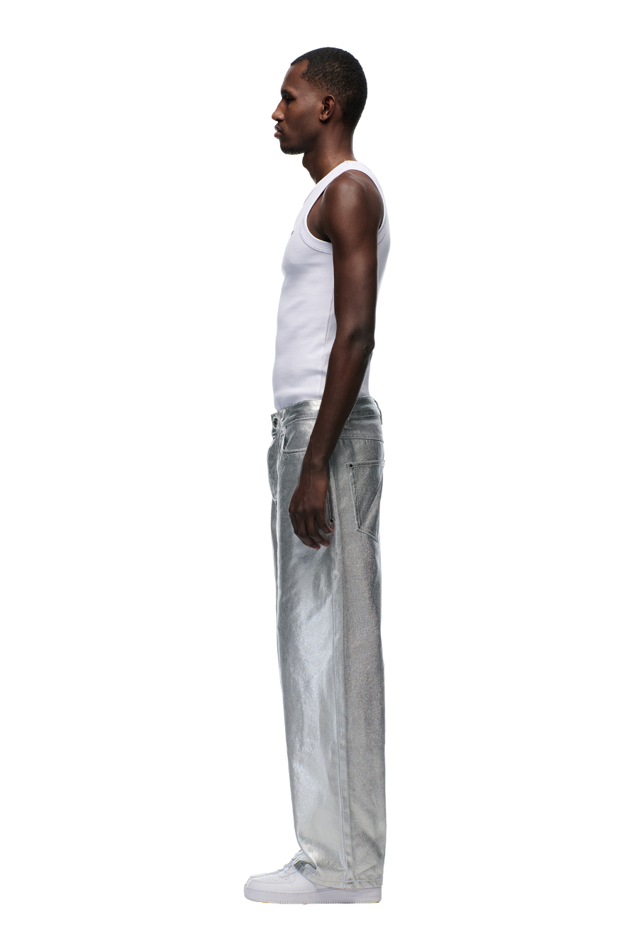 A man stands in profile against a white background, wearing a white tank top, 6PM’s WAX COATED DENIM SILVER SURFER pants, and white sneakers.