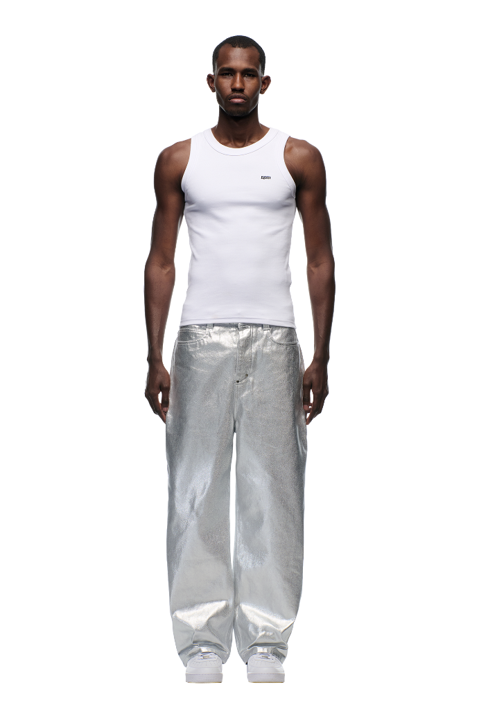 A man faces forward against a white background, wearing a fitted white tank, 6PM’s WAX COATED DENIM SILVER SURFER pants, and white sneakers.