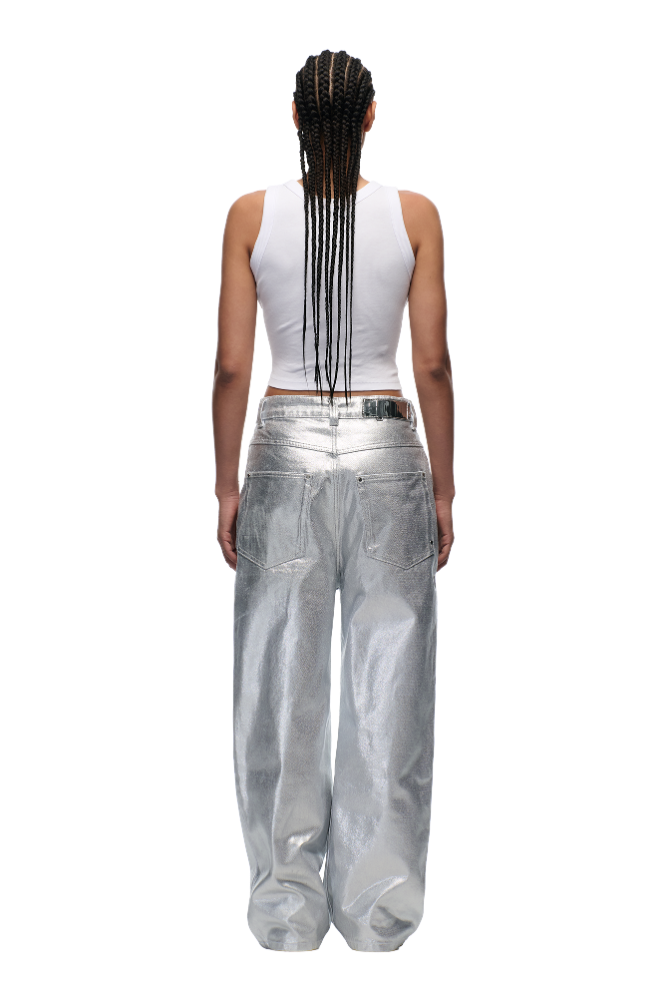 A person with long braided hair stands with their back to the camera against a plain white background, wearing a white sleeveless crop top and 6PM's WAX COATED DENIM SILVER SURFER baggy pants in shiny silver.