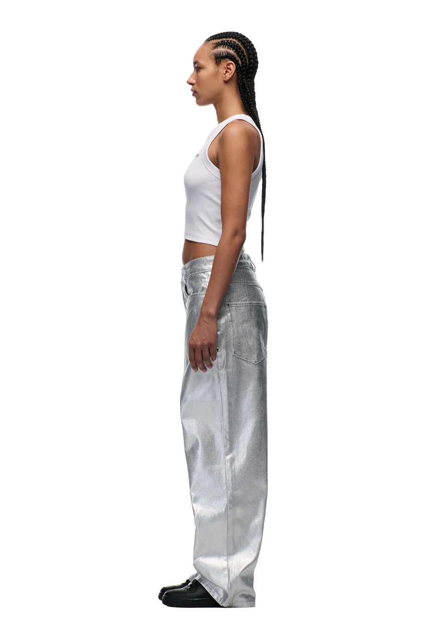 A woman with long braided hair wears a white sleeveless crop top, black shoes, and 6PM’s WAX COATED DENIM SILVER SURFER pants, standing in profile against a plain white background.