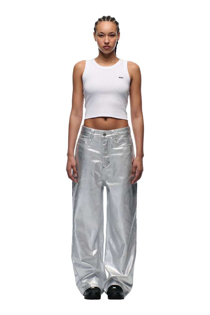 A woman with braided hair models the 6PM WAX COATED DENIM SILVER SURFER pants and a white crop tank top, standing against a plain white background with her arms at her sides and black shoes showing.