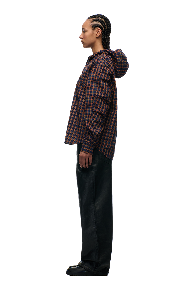A person stands in profile against a plain light background, wearing the 6PM HOODED DRESS SHIRT in blue, orange, and black checks with an adjustable hood, paired with black wide-leg pants and black shoes.