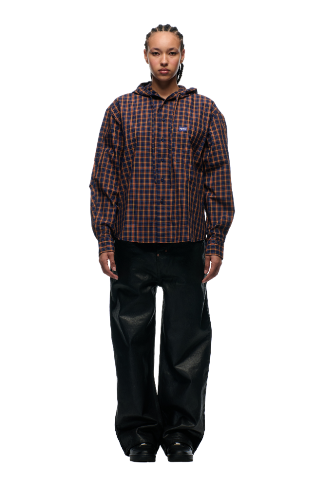 A person faces forward against a plain light background, wearing the 6PM HOODED DRESS SHIRT in blue, orange, and black checks with an adjustable hood, paired with loose black pants and black shoes.