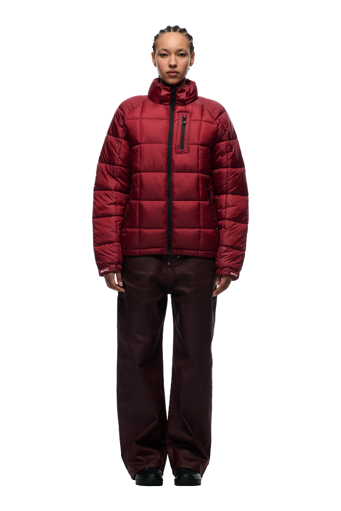 A person faces forward in the 6PM LIGHT PUFFER JACKET RECTANGLES WINE RED with a zippered chest pocket and adjustable hood, styled with dark wide-leg pants and black shoes against a white background—ideal for sporty 6PM looks.