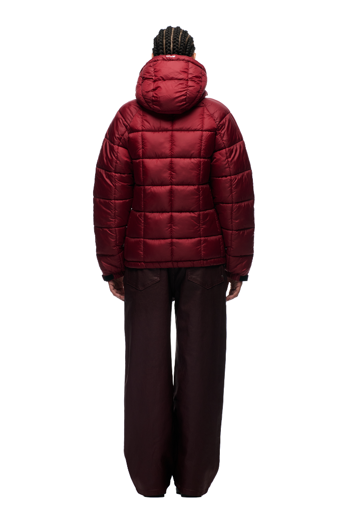 A person with braided hair faces away from the camera, wearing the 6PM LIGHT PUFFER JACKET RECTANGLES in wine red and loose dark pants, set against a plain white background.