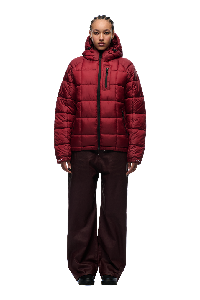A person faces forward against a plain light background, wearing the 6PM LIGHT PUFFER JACKET RECTANGLES WINE RED with an adjustable hood and chest pocket, paired with loose dark brown pants and black shoes.