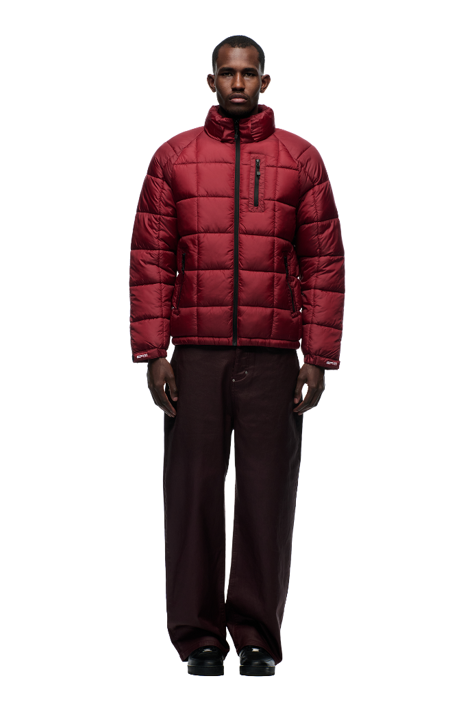 A man faces forward on a plain white background, wearing the 6PM LIGHT PUFFER JACKET RECTANGLES in wine red with a high collar and brown wide-leg pants.