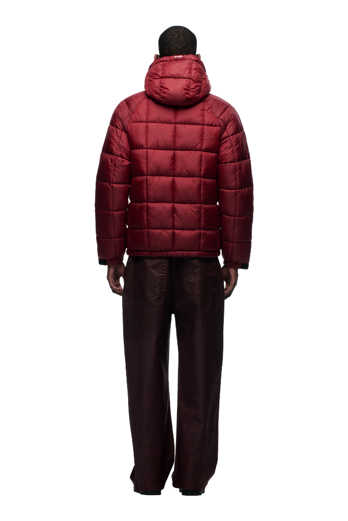 A person faces away, wearing the 6PM LIGHT PUFFER JACKET RECTANGLES WINE RED with an adjustable hood and loose dark brown pants, standing against a plain white background.