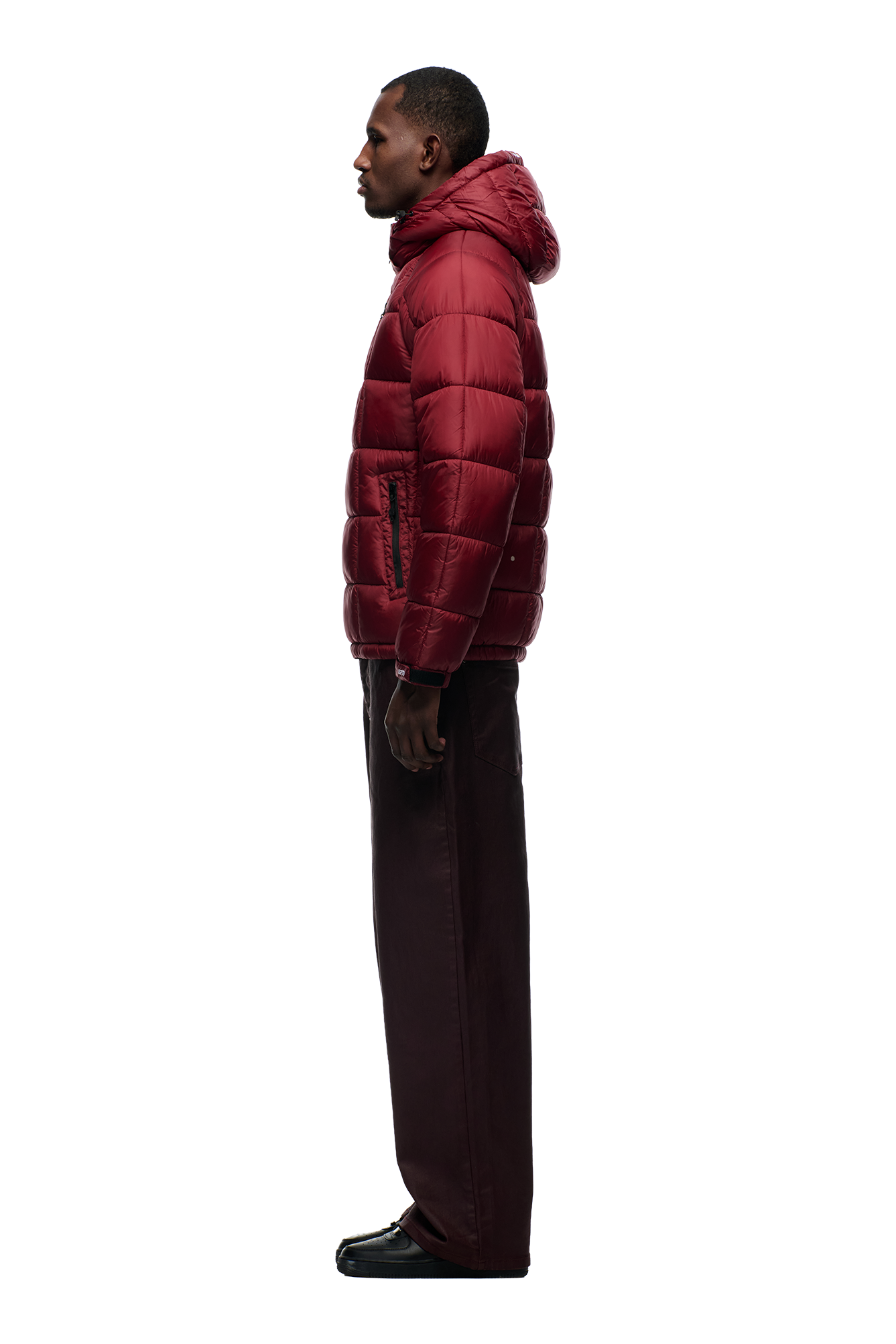 A person in profile wears the 6PM LIGHT PUFFER JACKET RECTANGLES WINE RED with an adjustable hood, paired with dark pants and black shoes, posed against a plain white background.