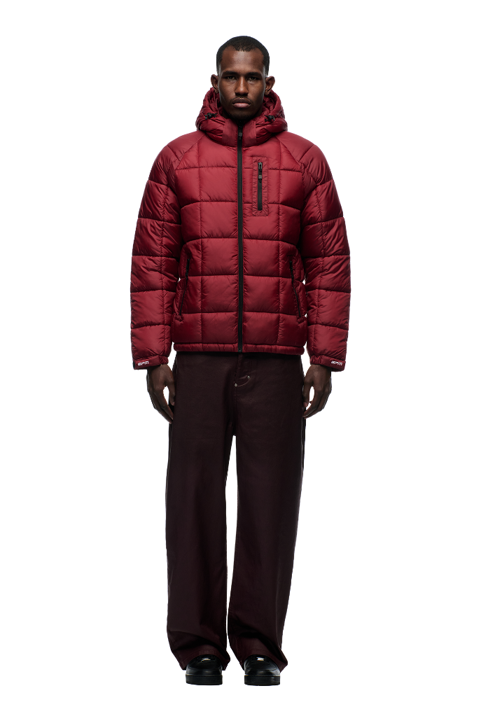 A person faces forward in a 6PM LIGHT PUFFER JACKET RECTANGLES WINE RED with an adjustable hood and zippered chest pocket, paired with loose dark brown pants and black shoes against a plain white background for a cozy sports-inspired look.