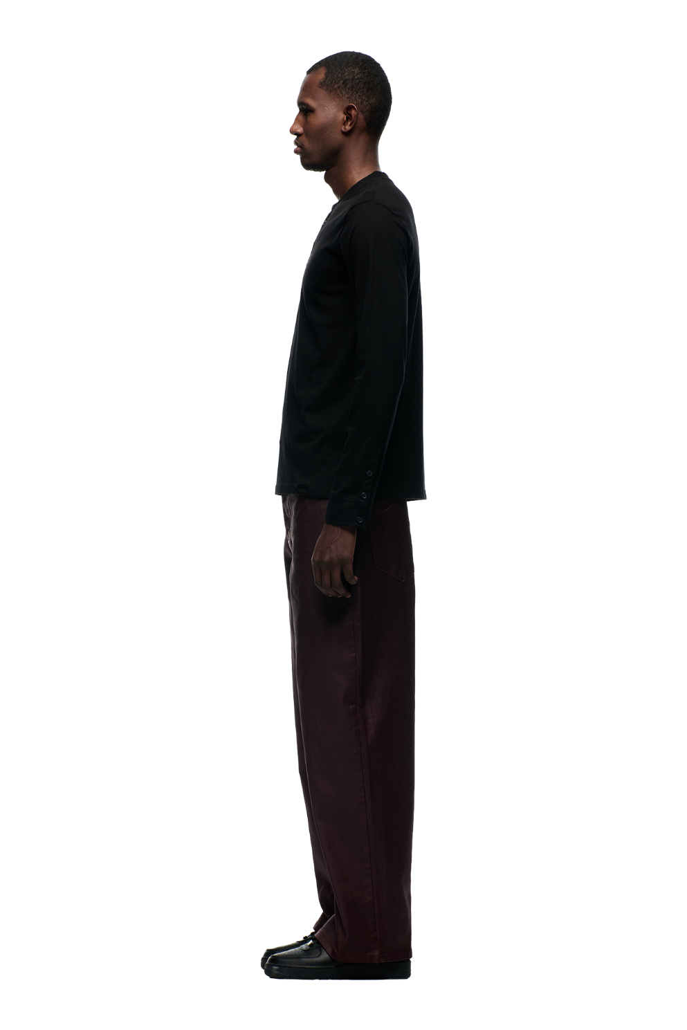 A man stands in profile against a plain white background, wearing the 6PM BUTTONED SLIM FIT LS BLACK shirt, loose dark brown pants, and black shoes.
