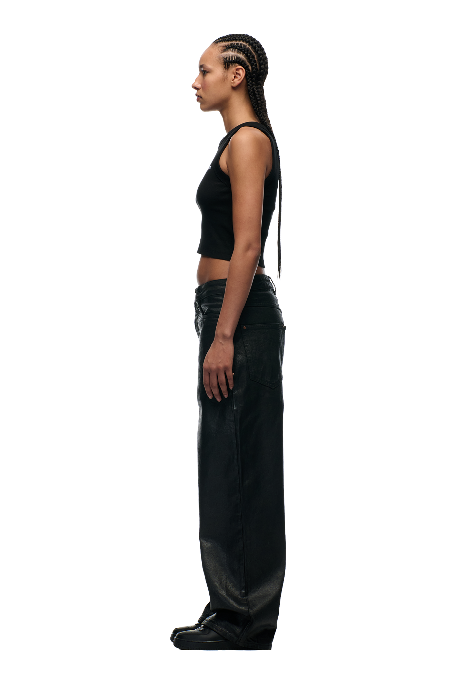 A woman with long braided hair stands in profile against a white background, wearing a black crop top, 6PM WAX COATED DENIM BLACK pants with metal buttons, and black shoes.