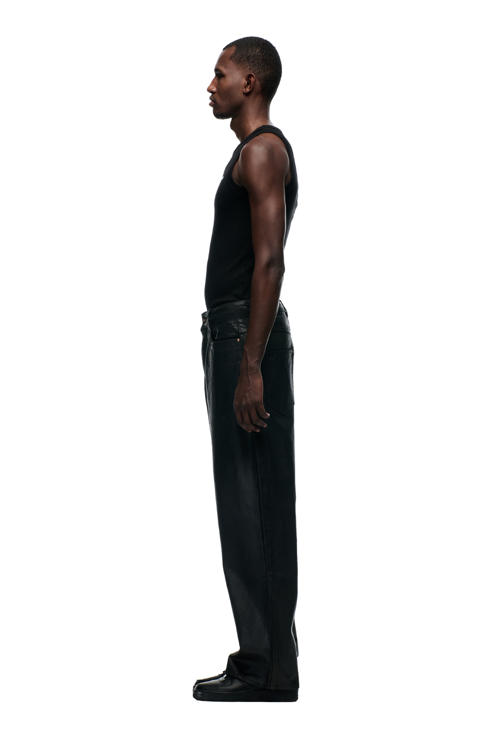A man stands in profile facing left, wearing a black sleeveless tank top, 6PM's WAX COATED DENIM BLACK pants with metal buttons, and black shoes, set against a plain white background.
