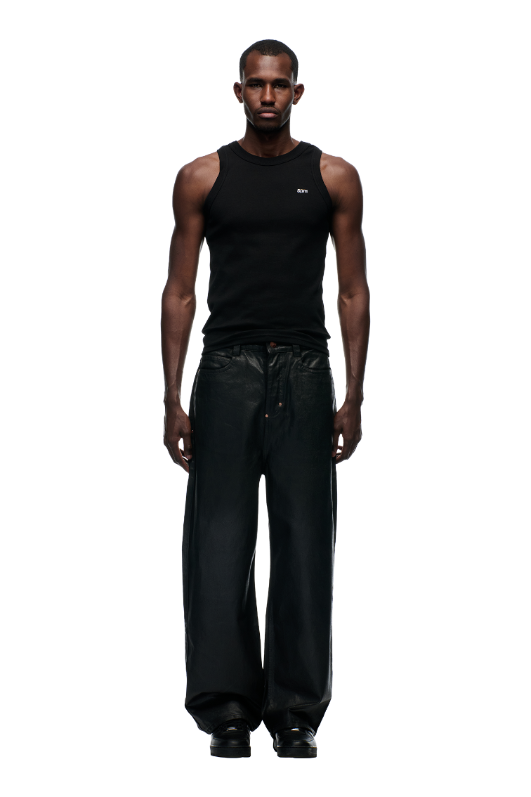 A man faces forward in a fitted black tank top and 6PM WAX COATED DENIM BLACK wide pants with drawstrings and metal buttons, styled with black shoes, set against a plain light gray background.