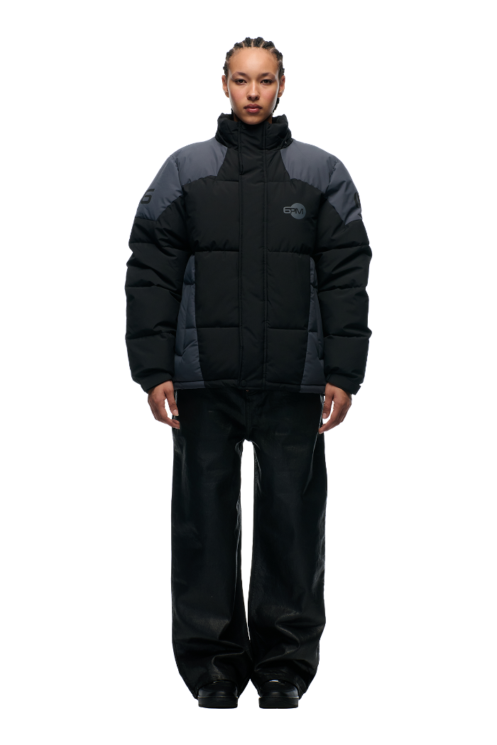 A person stands facing forward, dressed in the 6PM COLOUR BLOCKING PUFFER BLACK/ANTHRACITE jacket, paired with loose black pants and black shoes against a plain light background.