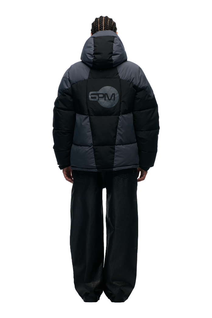 Person with braided hair wears the 6PM COLOUR BLOCKING PUFFER BLACK/ANTHRACITE with removable hood and bold 6PM logo on the back, paired with wide-leg black pants, facing away against a plain light background.