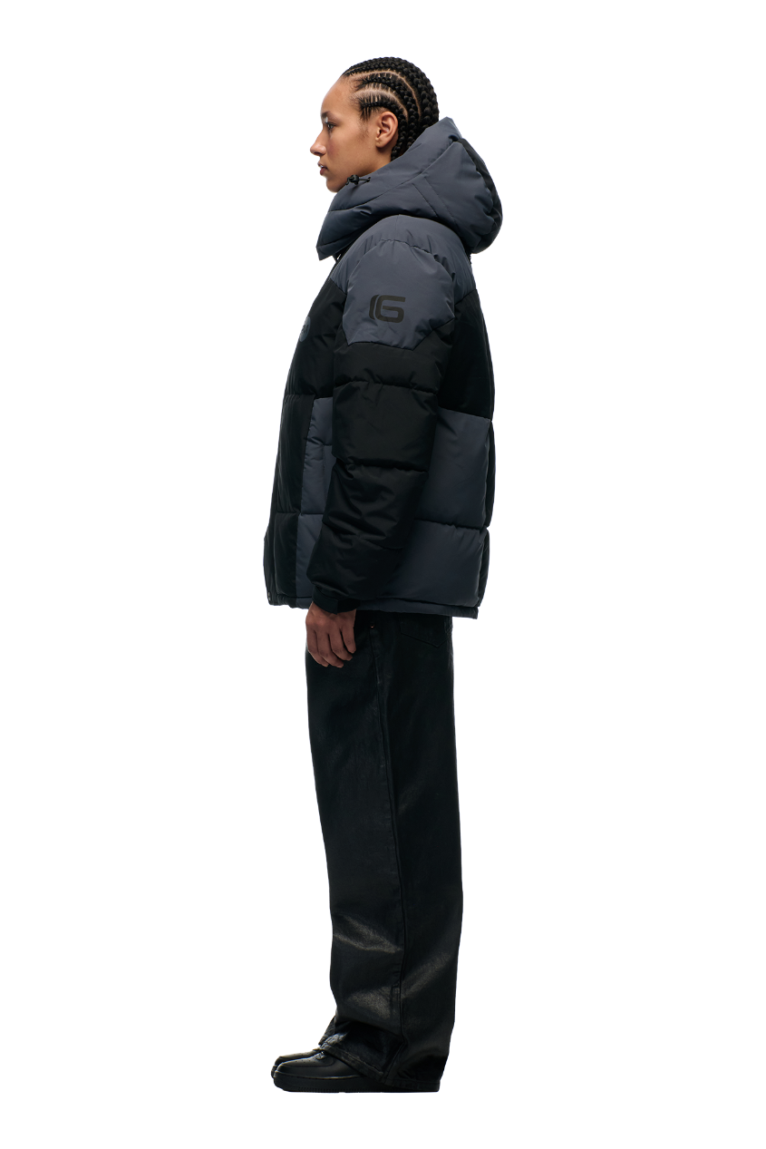 A person with braided hair stands in profile wearing the 6PM COLOUR BLOCKING PUFFER BLACK/ANTHRACITE with removable hood, loose black pants, and black shoes against a plain light gray background.