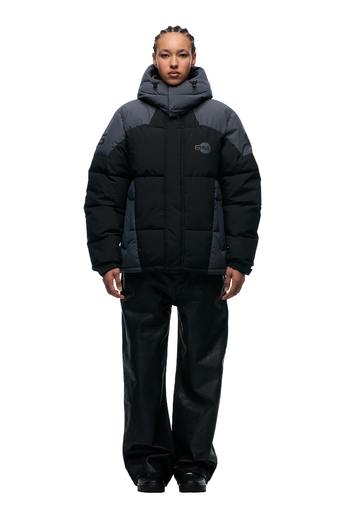 A person stands facing forward against a plain light background, wearing the 6PM COLOUR BLOCKING PUFFER BLACK/ANTHRACITE with removable hood, wide-leg black pants, and black shoes.