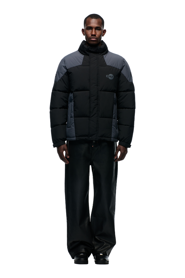 A man faces forward against a light gray background, wearing the 6PM COLOUR BLOCKING PUFFER BLACK/ANTHRACITE with a high collar and removable hood, paired with wide-leg black pants and black shoes.