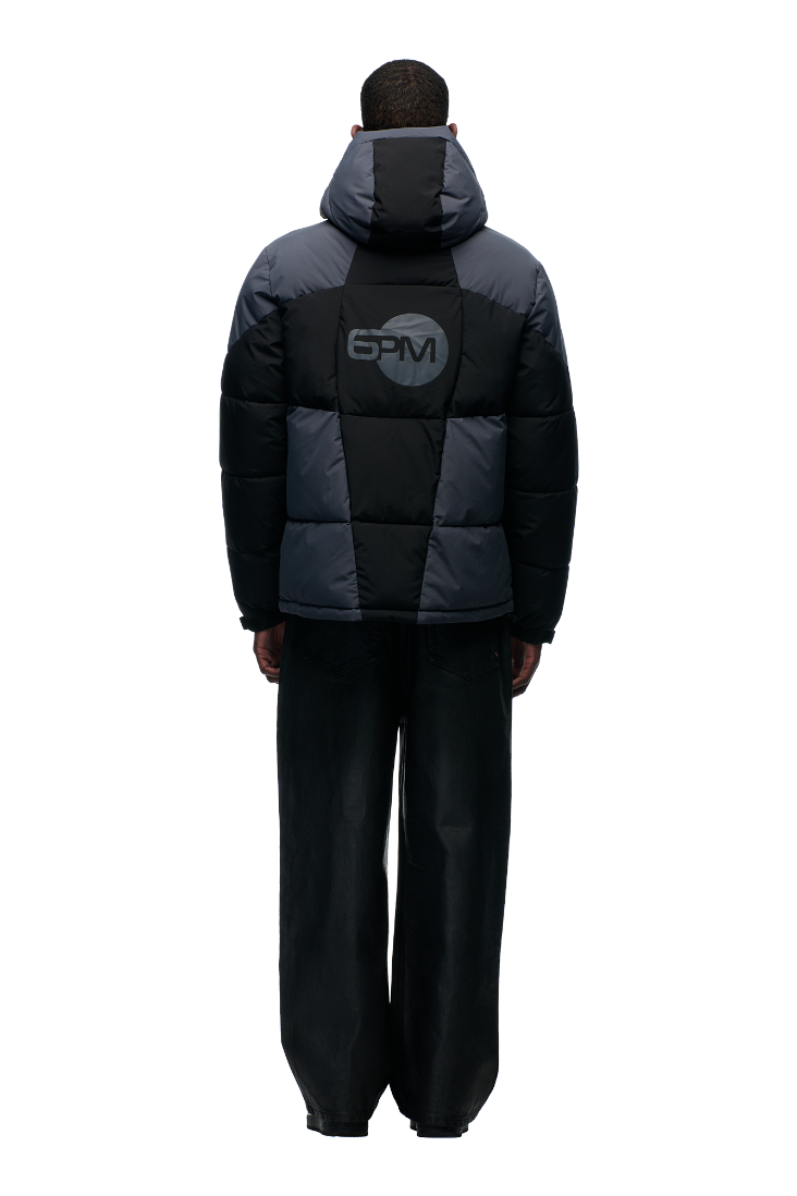 A person faces away, wearing the 6PM COLOUR BLOCKING PUFFER BLACK/ANTHRACITE jacket with a removable hood and a circular BPM logo on the back, paired with loose wide-leg black jeans. The background is plain white.