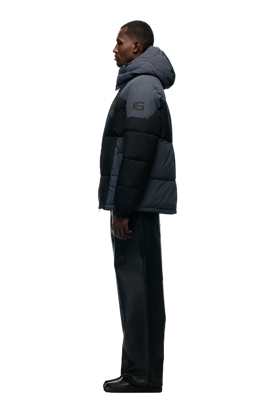 A man stands in profile against a light gray background, wearing the 6PM COLOUR BLOCKING PUFFER BLACK/ANTHRACITE with removable hood, black leather pants, and black shoes.