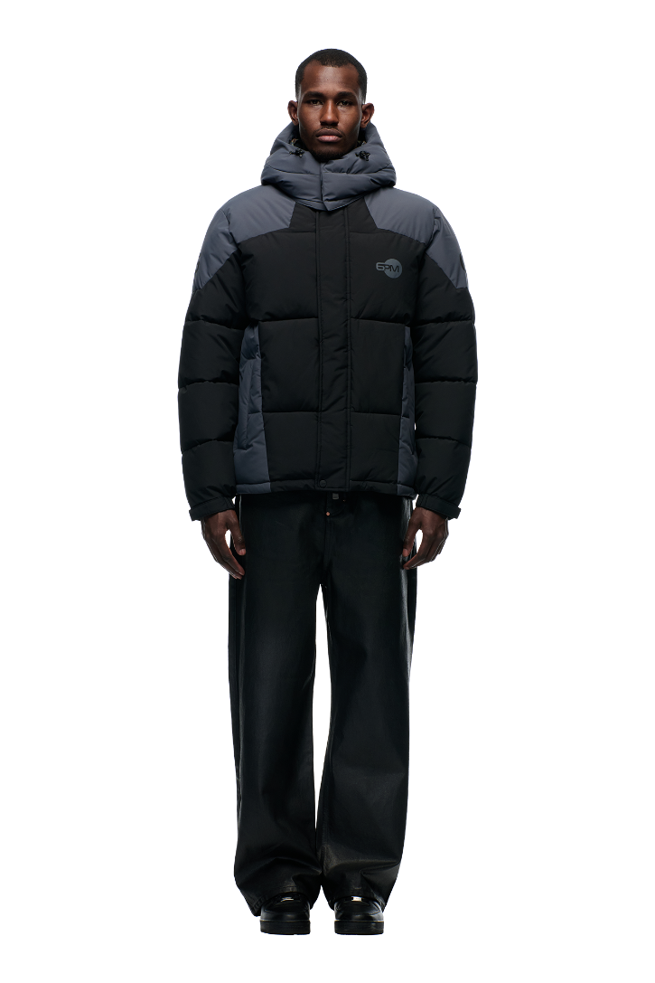 A man faces forward against a white background, wearing 6PM's COLOUR BLOCKING PUFFER BLACK/ANTHRACITE with a removable hood, wide-leg black pants, and black shoes.