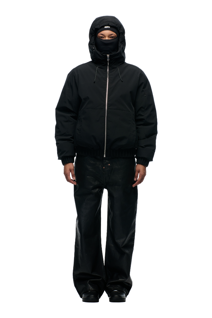 A person faces forward in a 6PM EMBOSSED JACKET BLACK, zipped up with the hood and high collar covering most of their face. The jacket features a hidden arm pocket. They wear matching loose black pants and shoes against a plain white background.