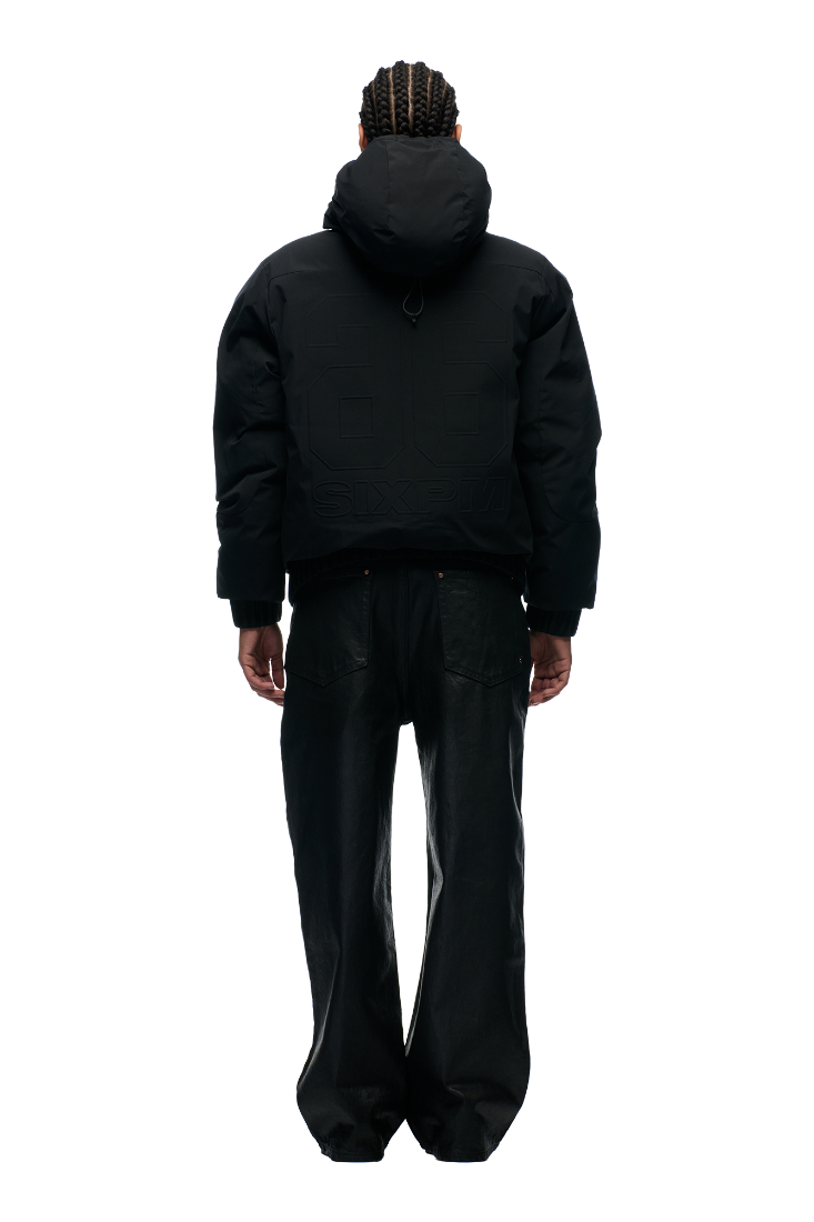 A person with braided hair, wearing the 6PM EMBOSSED JACKET BLACK with a hidden arm pocket and loose black pants, stands facing away from the camera against a plain white background.