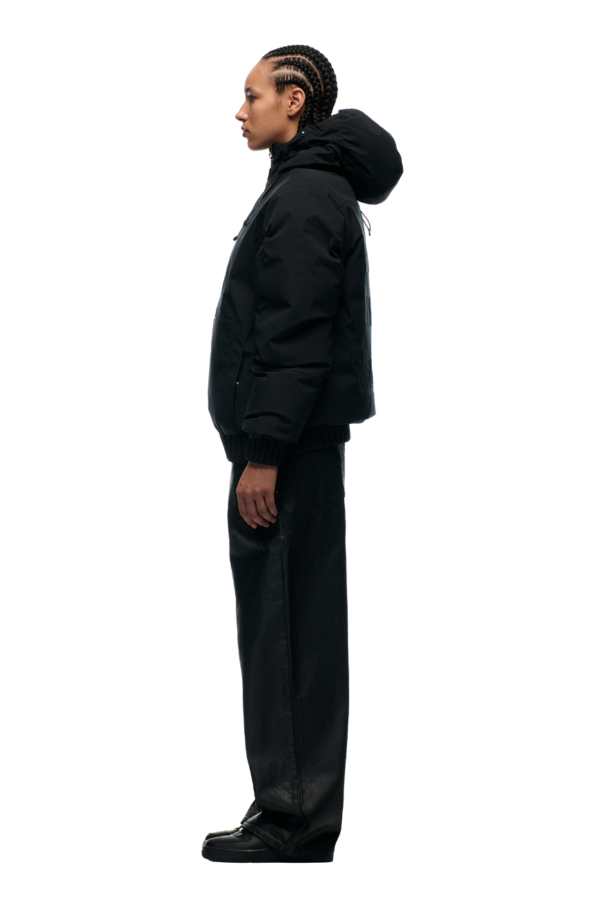 A person with braided hair stands in profile against a plain white background, wearing the 6PM EMBOSSED JACKET BLACK, paired with wide-leg black pants and black shoes.