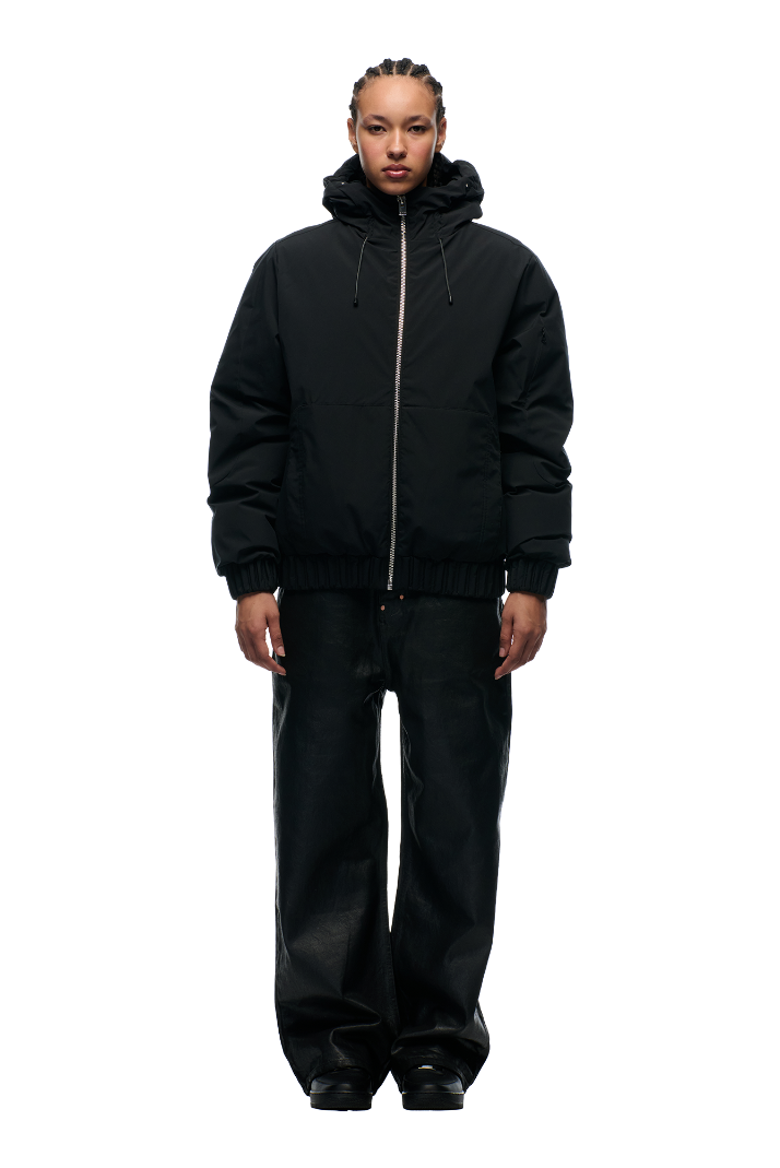 A person with braided hair wears the 6PM EMBOSSED JACKET BLACK with a hidden arm pocket, paired with loose black pants and black shoes, standing facing forward against a plain white background.