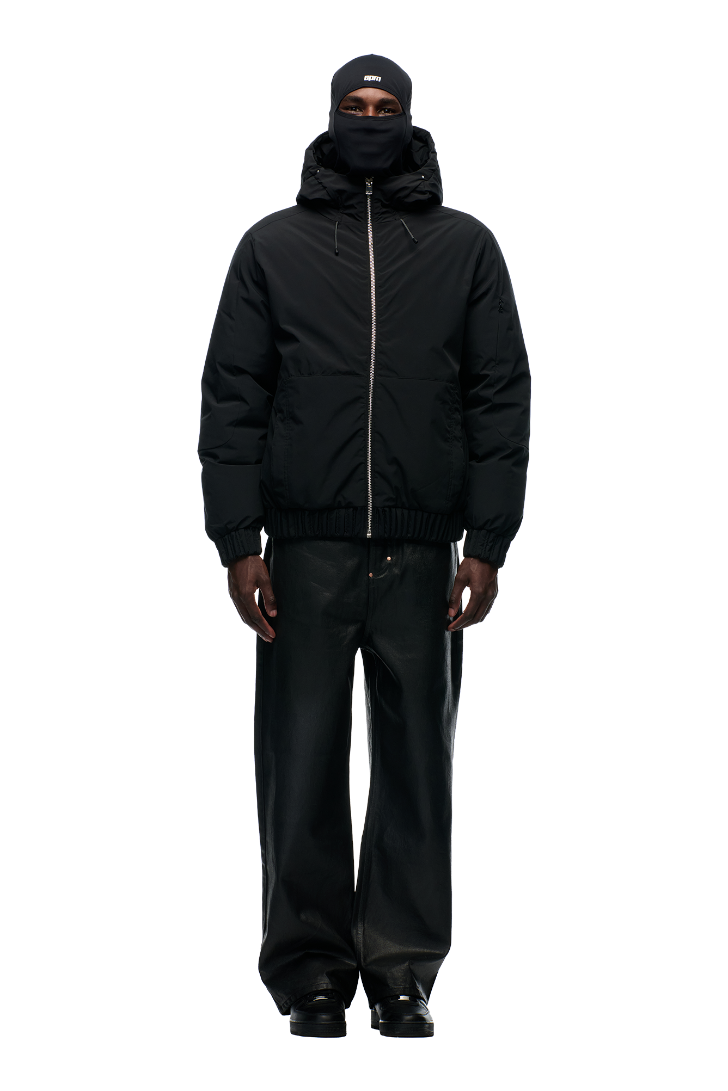 A person stands facing forward in the 6PM EMBOSSED JACKET BLACK, featuring a hidden arm pocket, paired with a black balaclava, loose black pants, and black shoes against a plain light background.