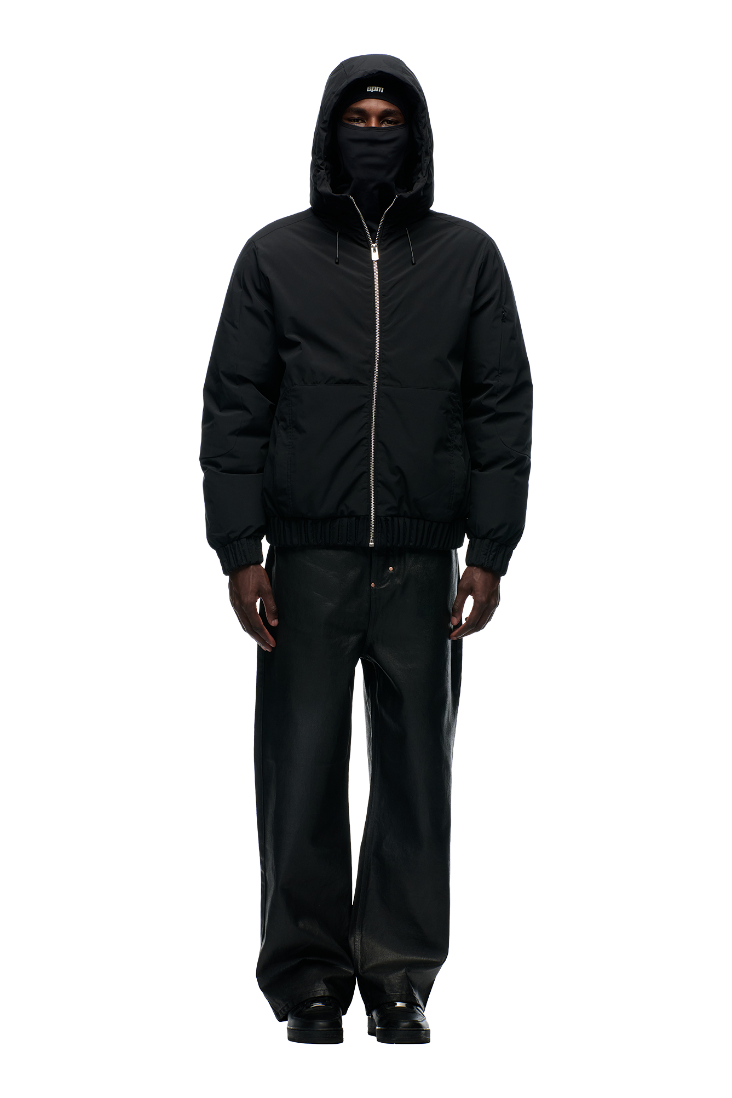 A person stands facing forward wearing the 6PM EMBOSSED JACKET BLACK, featuring a reflective cord, paired with a black face covering, wide-leg black pants, and black shoes against a plain light gray background.